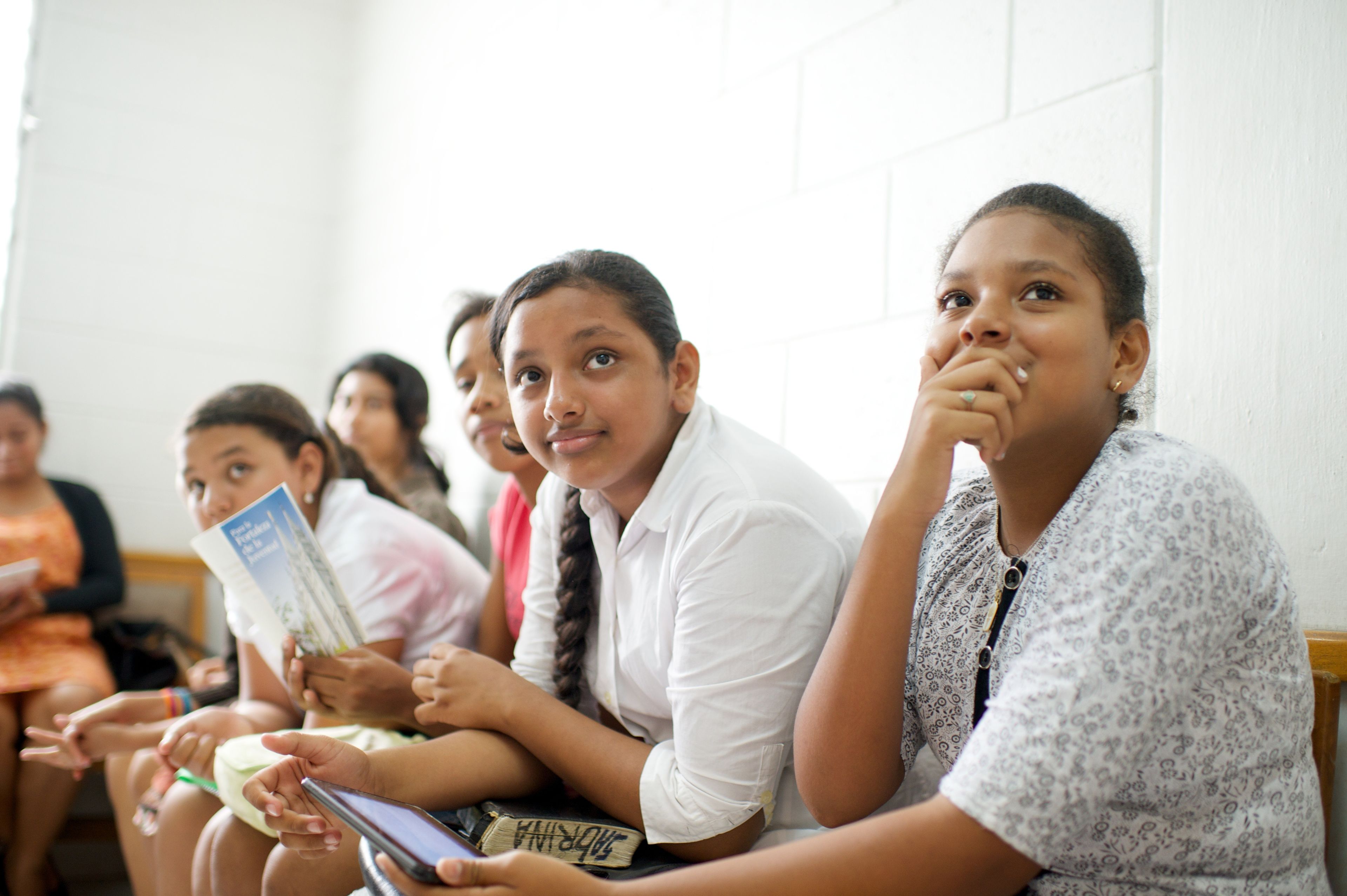 Young women from Ecuador sitting in a row at church, with one reading For the Strength of Youth.