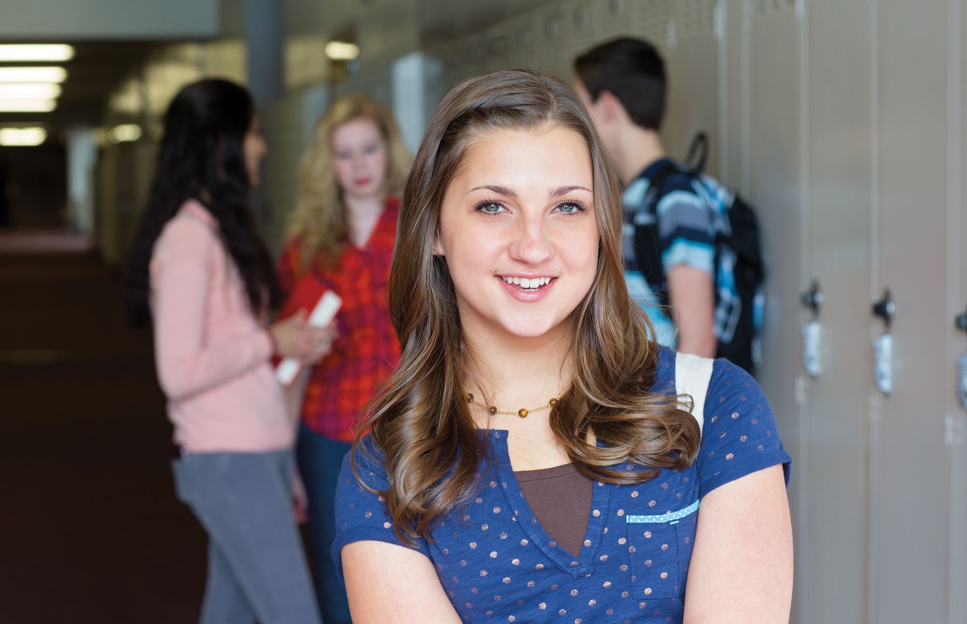 A portrait of a young woman standing near her locker in a high school, with other teenagers in the background.