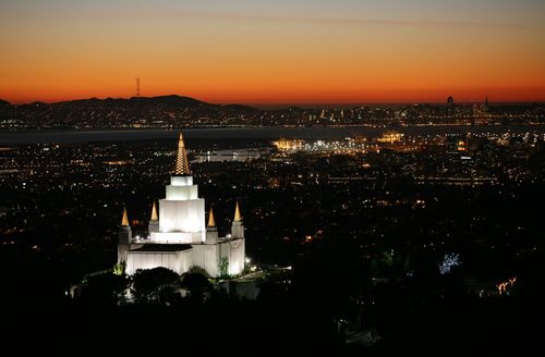 The Oakland California Temple illuminated in the evening, with the lights of the city showing in the background.