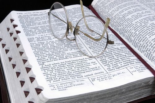 A pair of rounded wire-frame eyeglasses lying on top of a Bible, which is opened to Acts chapter 27 and marked with a maroon ribbon.