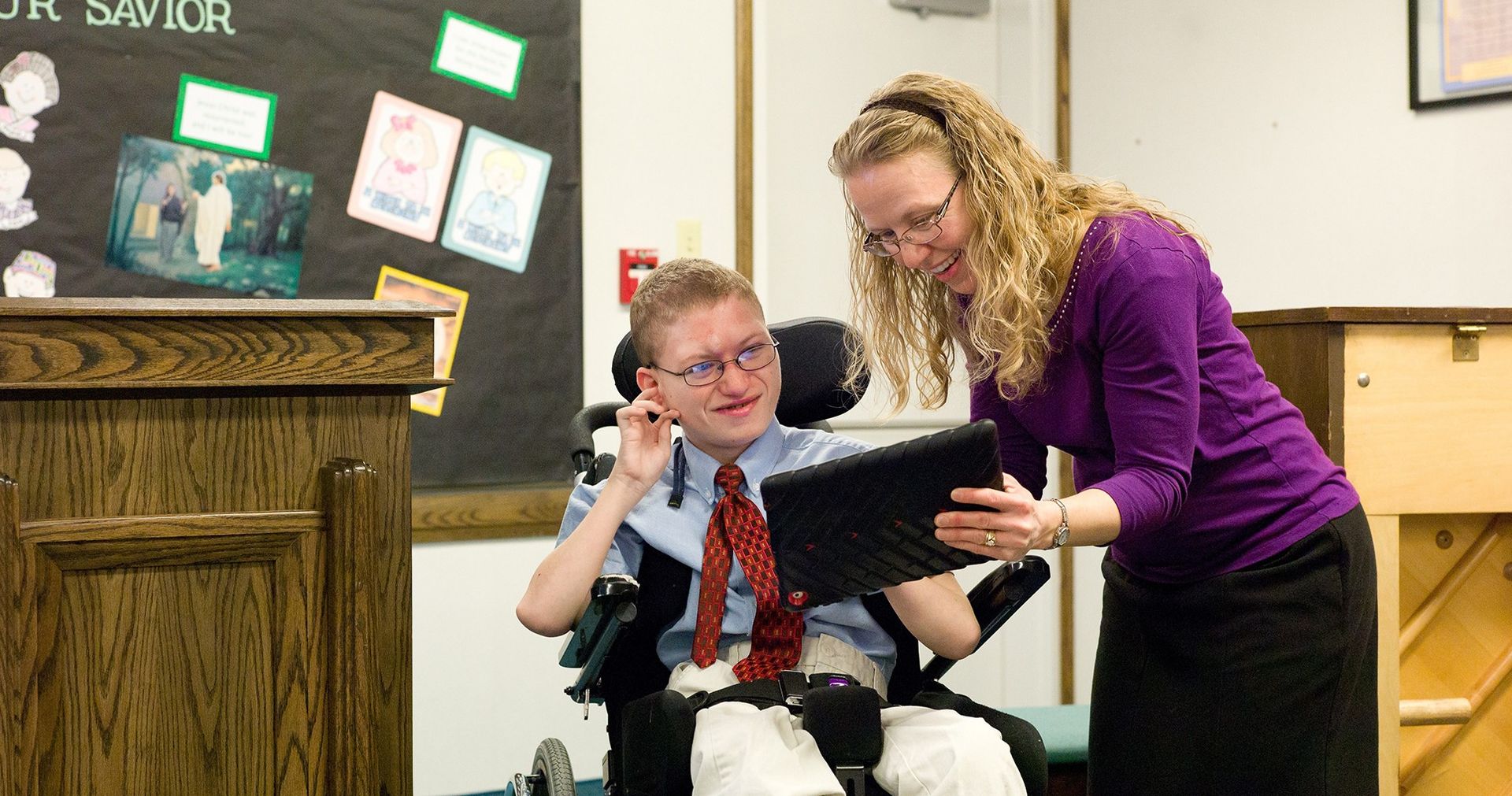 Teacher helping a young man in a wheelchair present at the front of a class.