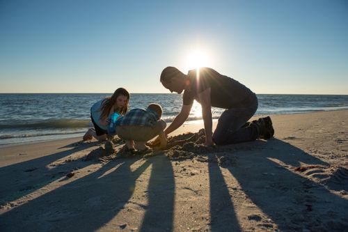 A man in a T-shirt kneels in the sand with his two children to build a sand castle together on a beach at sunset.