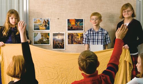 A teacher and children standing in front of a group of children.  They are lowering a blanket to reveal pictures on the bulletin board.