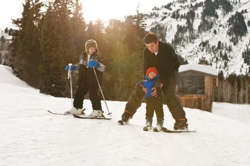 A father on skis helps push his son on skis while his other son stands next to them with skis and poles.