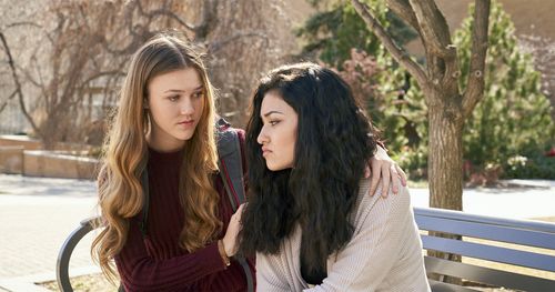 Two young women sit together on a bench. One of the young women appears to be distraught, sad, or concerned about something. The other young woman is showing concern and appears to be trying to comfort the other.