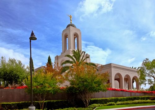 The front and part of the side of the Newport Beach California Temple, with the spire rising over nearby trees and a blue sky overhead.