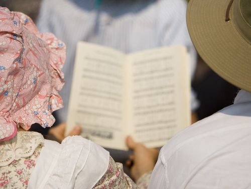 A woman wearing a bonnet and a flower-patterned dress holds a hymnbook next to a man in a white shirt and wide-brimmed hat.