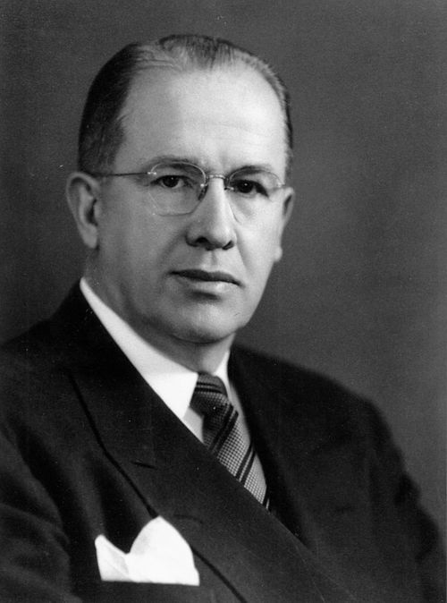 President Ezra Taft Benson in a white shirt, striped tie, and black suit with a handkerchief in his front pocket, around 1949.