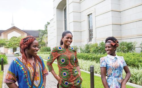 Three women in colorful clothing, smiling as they walk on the grounds of the Accra Ghana Temple.