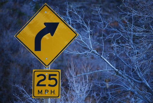 A yellow road sign with a curving black arrow and the marking “25 M.P.H.” standing near bare trees in the wintertime.