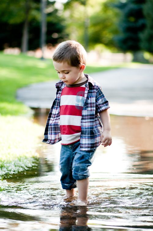 A toddler boy with rolled-up pants walks outside in a deep puddle of water.