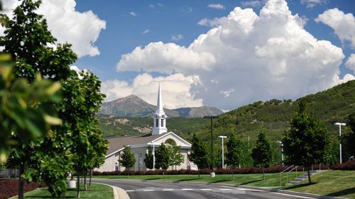 Green, leafy trees and a road leading to a tan chapel with a white steeple and mountains in the background in Draper, Utah.