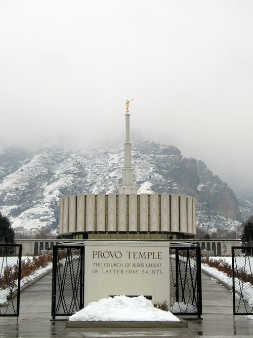 A front view of the Provo Utah Temple on a gray winter day, with the temple’s sign on the black fence and the gates open leading to the temple.