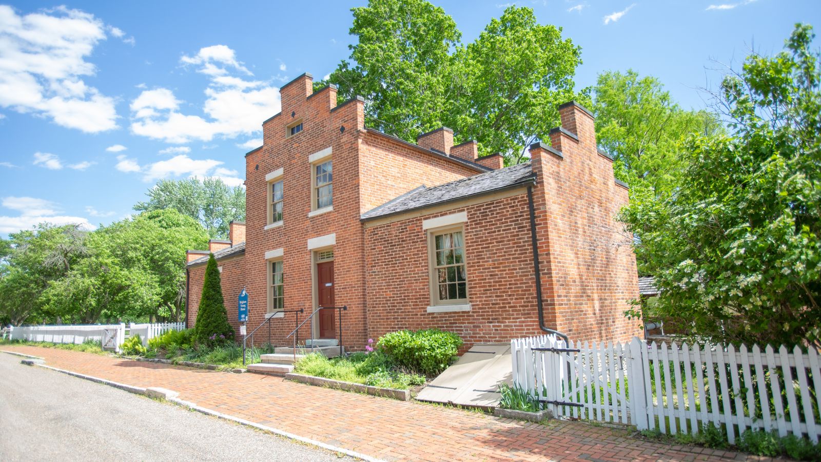 A large one and a half story red brick house. A white fence surrounds it, and large green trees stand behind it. 