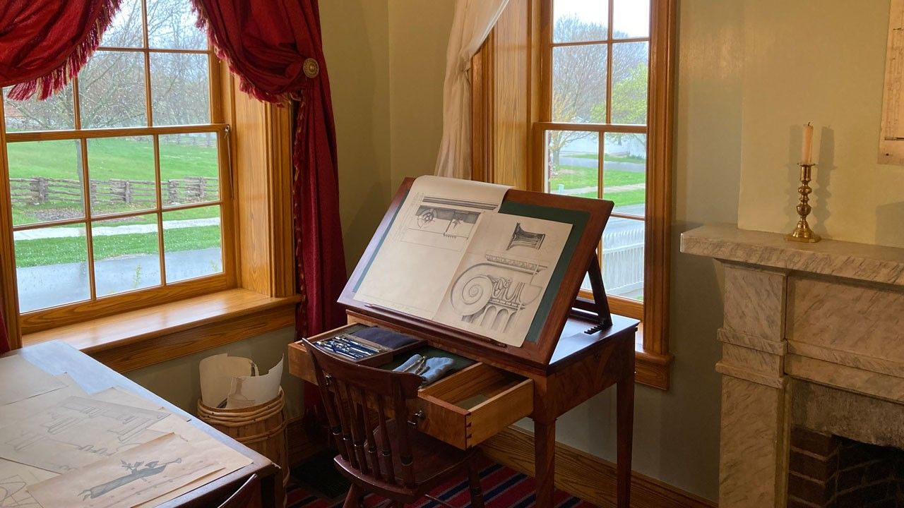 A tilt-top desk with architectural drawings on it in the corner of a small room with windows offering views outside. 