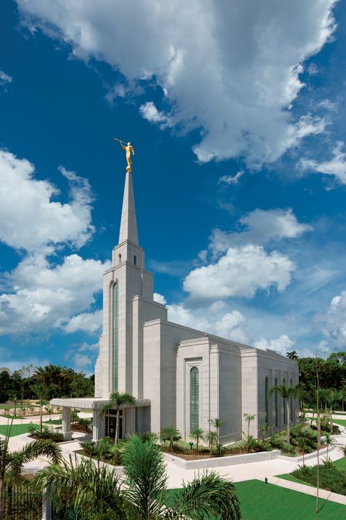 A view of the Manaus Brazil Temple at an angle from afar, showing the surrounding landscape on a sunny day with a cloudy blue sky.