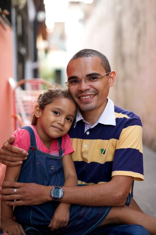 A man in a blue and yellow striped shirt holds his daughter, who is in a denim jumper, while the two of them smile at the camera.