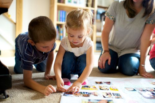A boy and a girl kneel down in a living room by their mother and make a family tree with family photos.