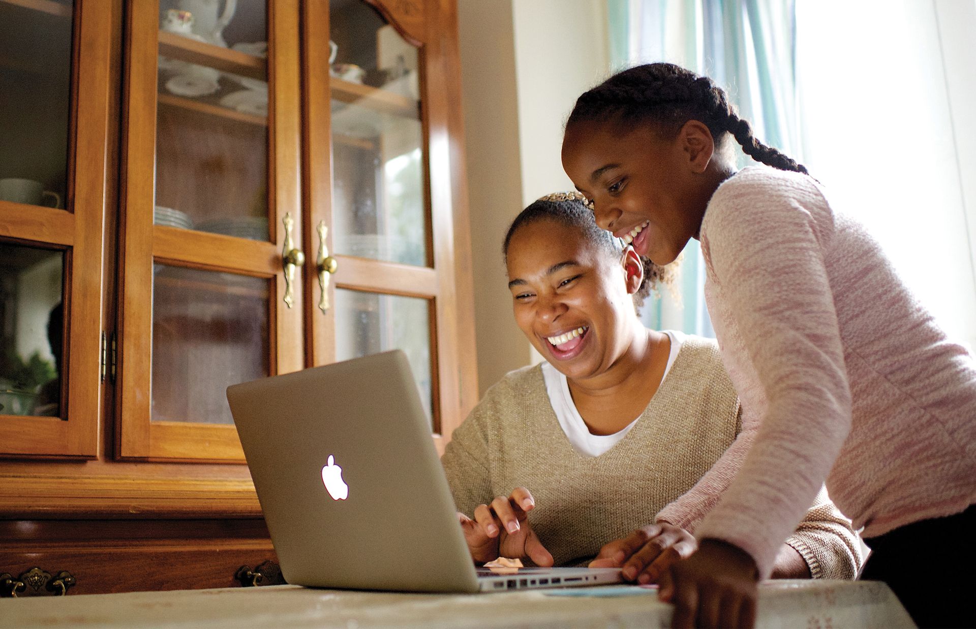 A mother and daughter work on a computer together in the dining room.