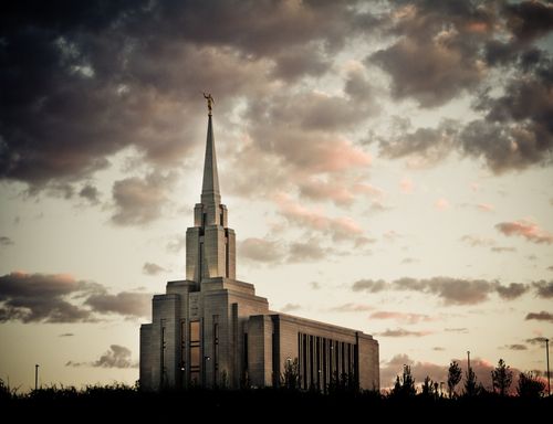 A front side view of the Oquirrh Mountain Utah Temple in the evening, with the temple’s lights on and purple clouds overhead.