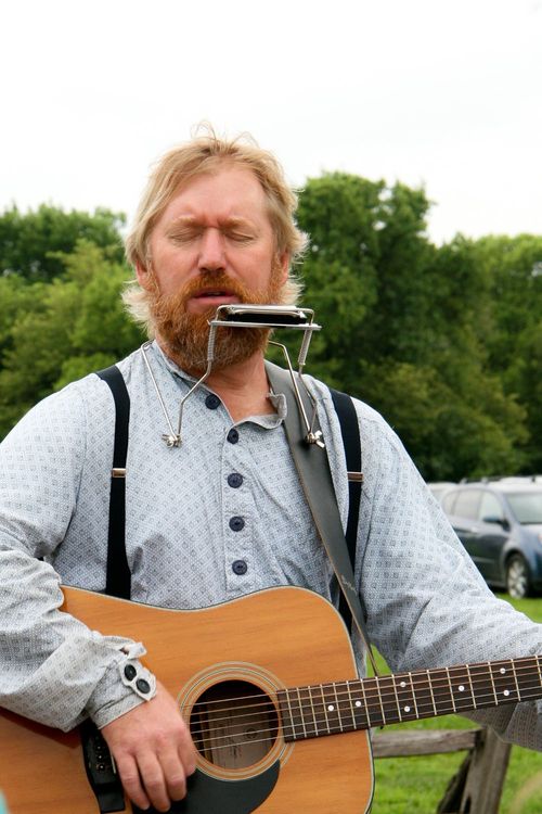A man standing outside at the Nauvoo Pageant, playing his guitar and singing with a harmonica attached to his shirt, ready to play.