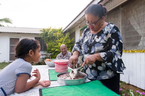 A woman stands at an outdoor table cutting a round cake and interacting with her granddaughter.