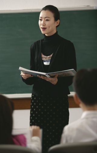 A Korean woman standing in front of a group of Primary children.  She is holding a songbook.