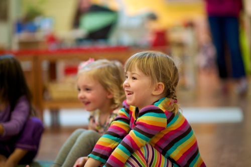 A little girl with pigtails and rainbow-striped clothing sits next to another little girl on the floor in a classroom at an elementary school.