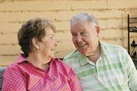 An older couple sitting together outdoors on a bench.  They are smiling and laughing.