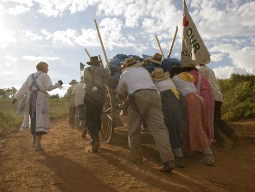 A man and three women dressed as pioneers push a handcart full of equipment while others stand on the side and help guide it over a dusty path.
