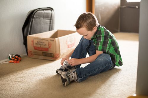 A young boy with brown hair and a green plaid shirt sits on the carpet to tie up his shoelaces.