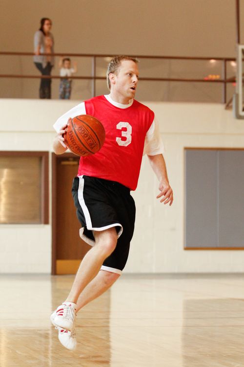 A man in a red jersey, black shorts, and tennis shoes plays basketball at a church gym, with his wife and child watching from above.