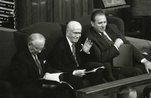 President Benson sitting in a large armchair between his counselors, smiling and waving to the audience in the Tabernacle.