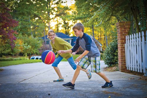 A boy dribbles a basketball with his sister coming up behind him as they play in the driveway.