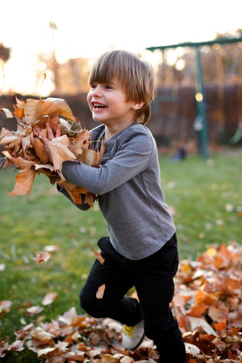 A young boy is playing outside, running with a pile of leaves in his hands and a smile on his face.