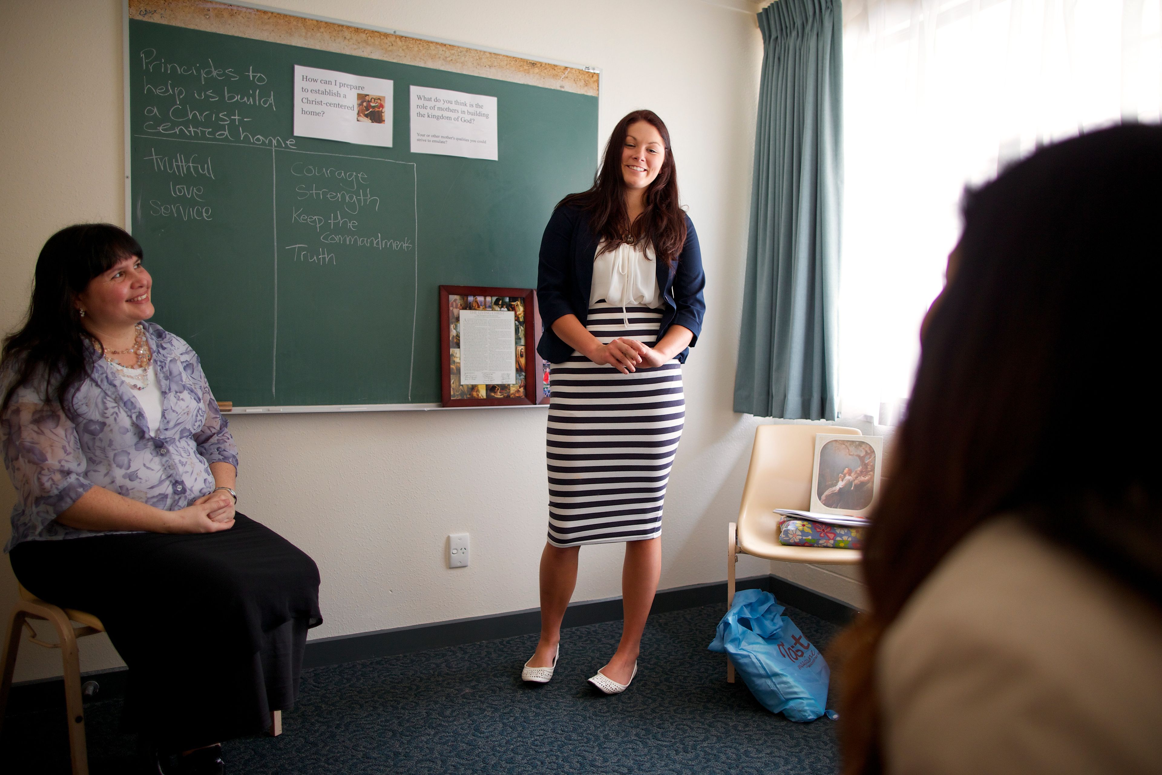 A young woman stands up in front of her class and teaches a lesson with her teacher sitting nearby.  