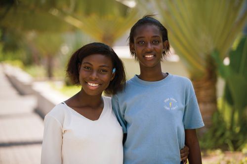 A portrait of two young women smiling and standing outside with their arms around one another.