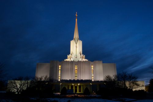 The front of the Jordan River Utah Temple on a winter night, with yellow light shining through the windows and bare trees near the entrance.