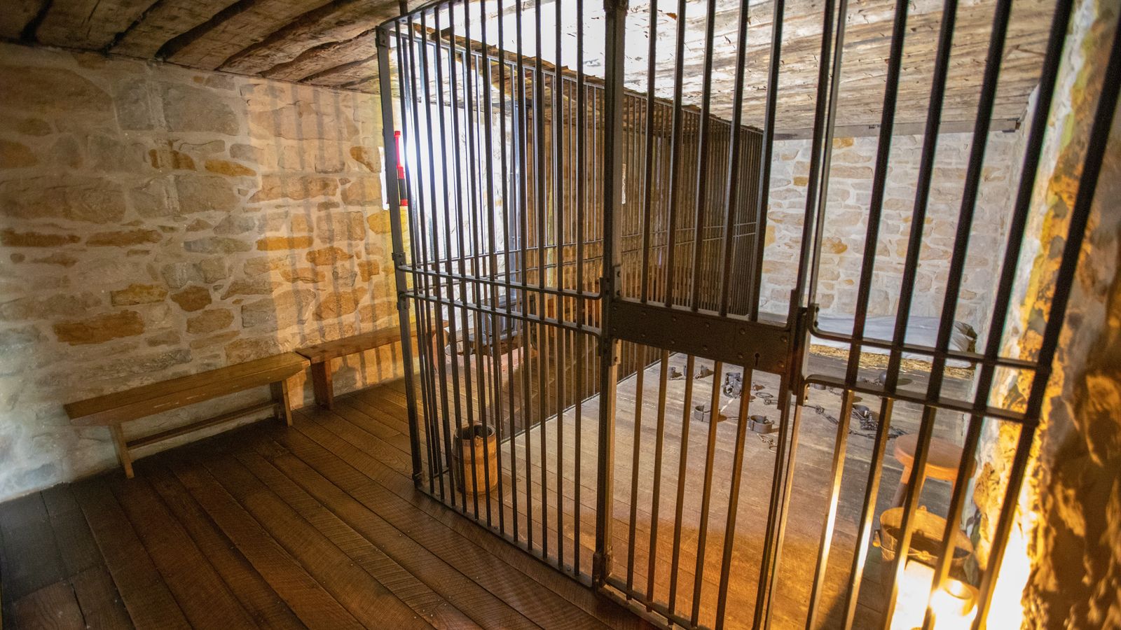 A small room with stone walls and a floor-to-ceiling cage made of metal bars. Light from a lantern illuminates the room.