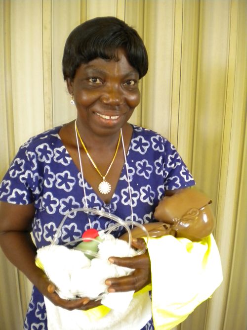 A Ghana midwife in a blue and white floral dress standing and holding a CPR practice baby wrapped inside a blanket.