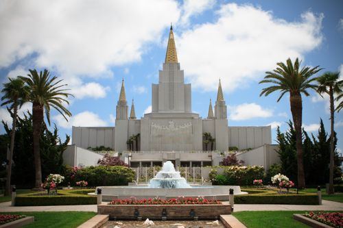 A view of the front of the Oakland California Temple, with palm trees and flowers growing on the grounds and a large water fountain in view.