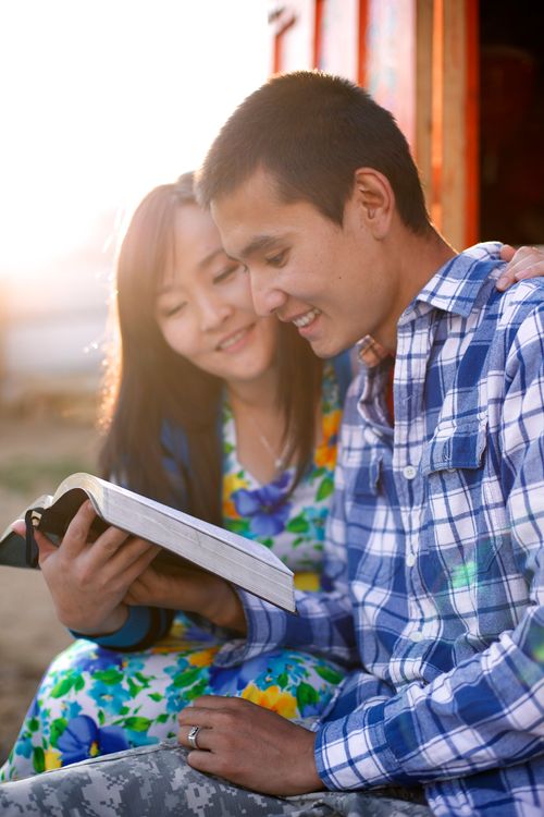 A woman in a floral dress sits next to a man in a plaid shirt as they read the scriptures together outside in Mongolia.