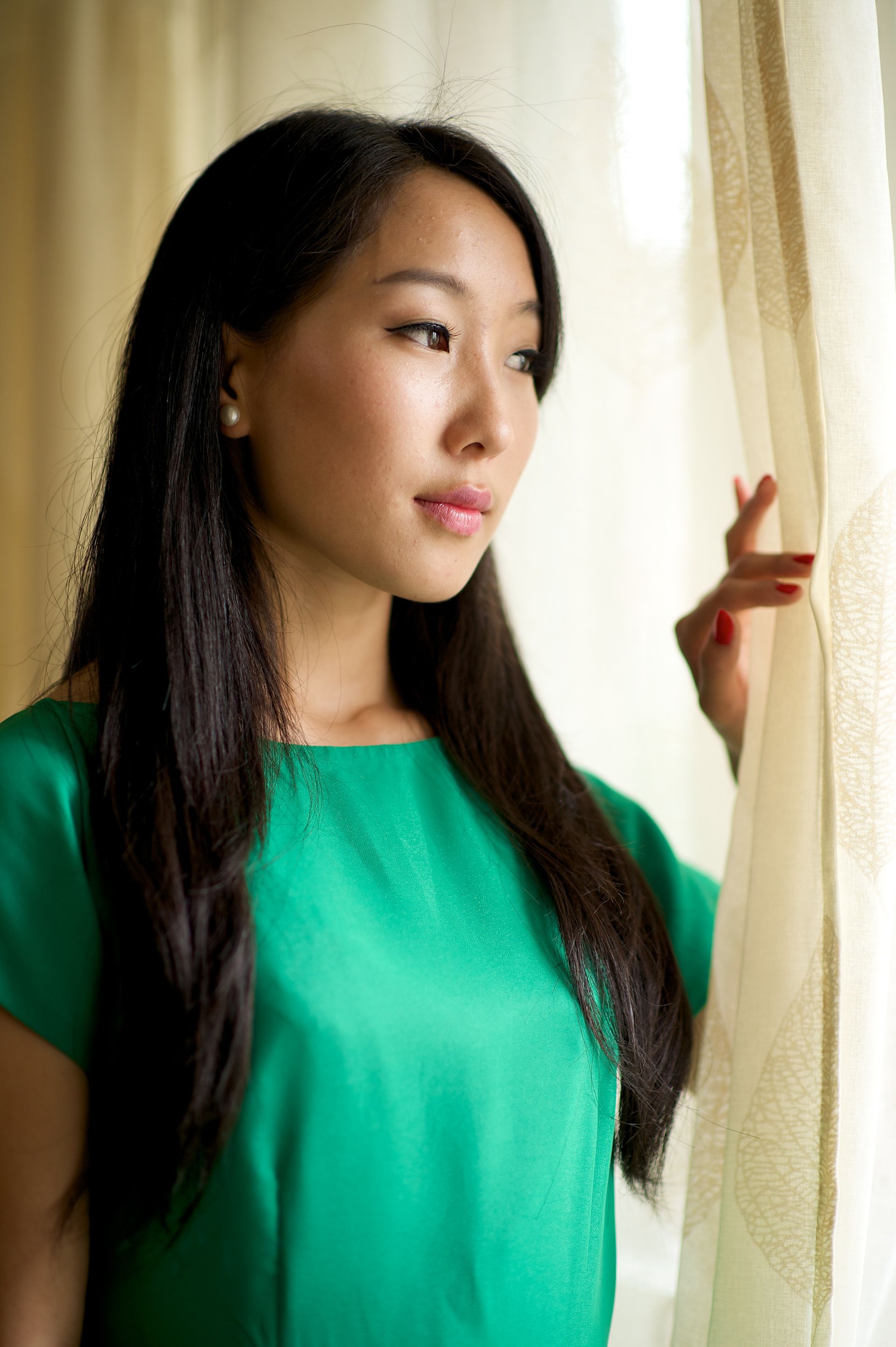 A young woman in Mongolia holds a curtain open and looks out a window.