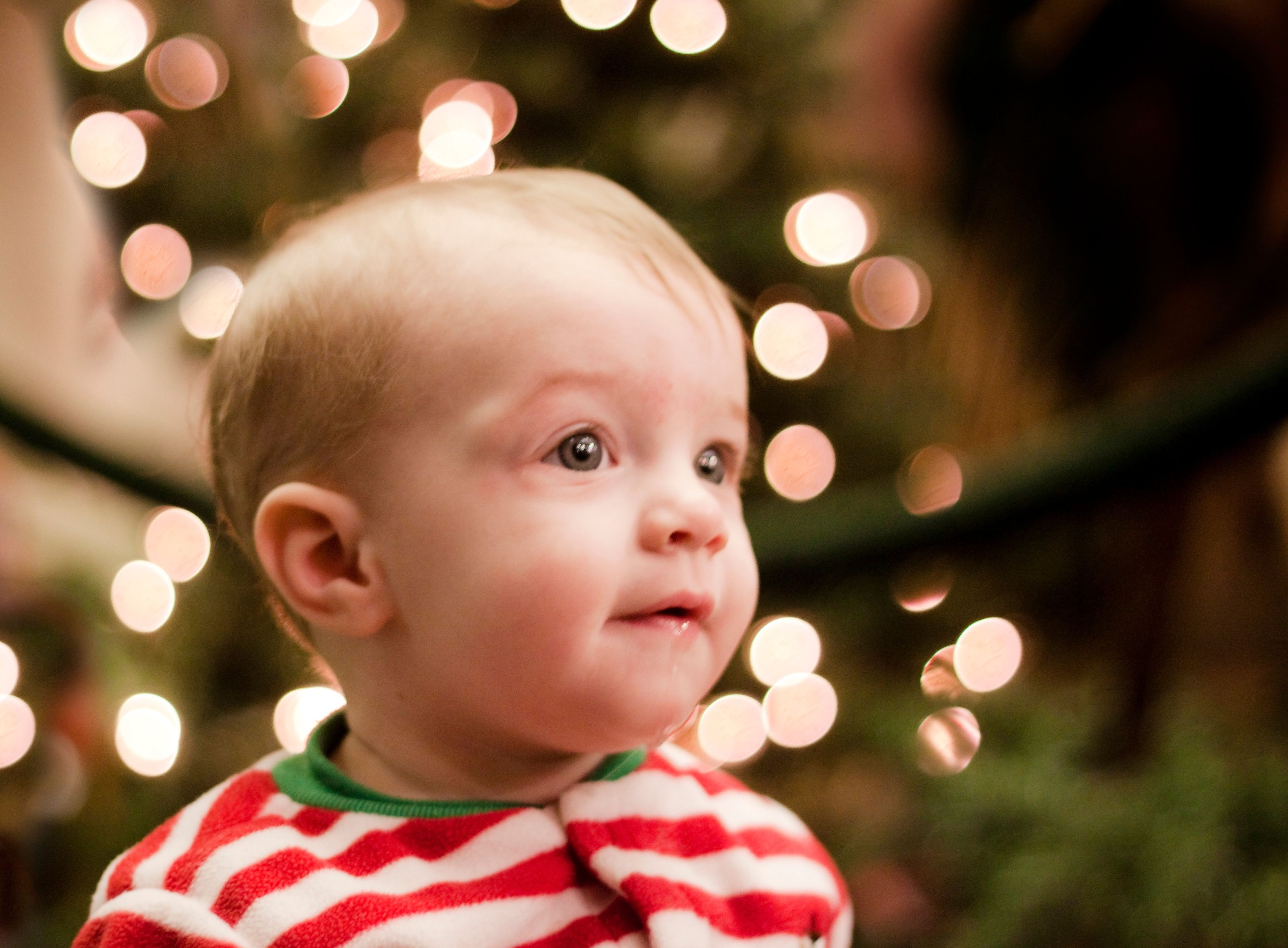 A baby in red, white, and green pajamas sitting near a Christmas tree.