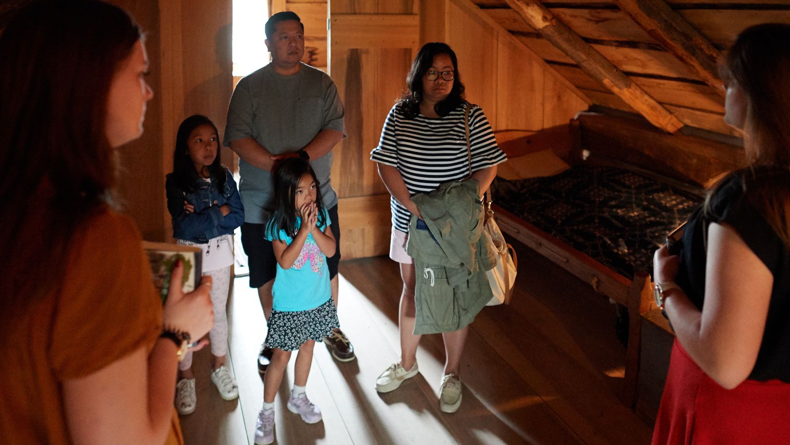 An interior view of Joseph Smith's family home in Palmyra, New York. 
A family walks inside and interacts with various setups inside. They see artifacts within the home. They talk with Sister Missionaries inside the home.