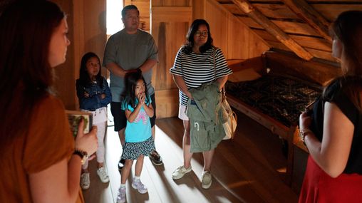 An interior view of Joseph Smith's family home in Palmyra, New York. 
A family walks inside and interacts with various setups inside. They see artifacts within the home. They talk with Sister Missionaries inside the home.