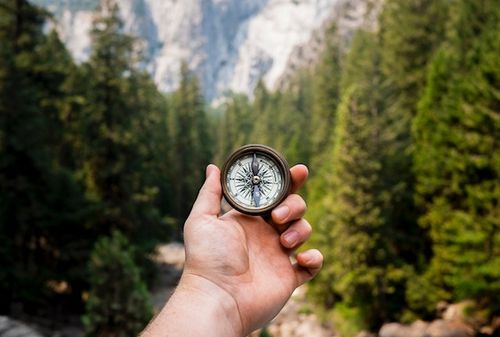 A hand is holding up a compass. You can see mountains and trees in the background.