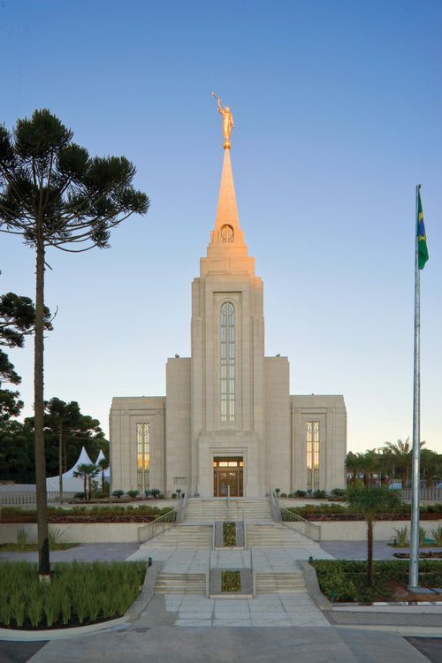 The Curitiba Brazil Temple in the evening, with a set of stairs leading to the front entrance and the Brazilian flag nearby.
