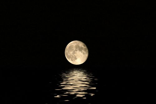 A full moon in a pitch-black sky reflects on the water.