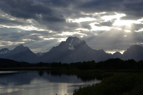 Dark clouds gather over the Teton mountain range with sun rays breaking through and shining down over the mountains and lake.